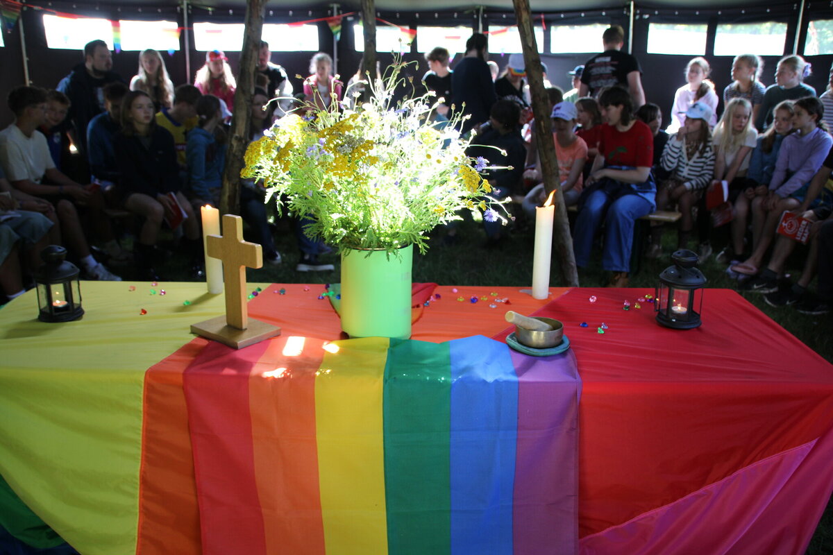 Andacht mit Regenbogen-Tischdecke, Kerzen, Blumen und Kreuz im Zelt